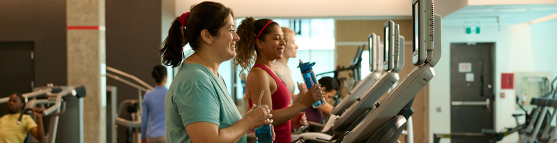 Two people jogging side by side on treadmills at a gym, smiling and holding water bottles, with other people exercising in the background.