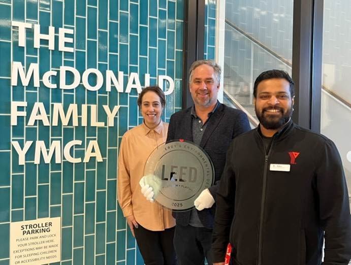 Three adults stand in front of a teal tiled wall with the words The McDonald Family YMCA in large white letters. From left to right: a woman with dark hair pulled back wearing a beige blouse (Sarah Beldick); a man with short gray hair and beard wearing a dark blazer and holding a round glass LEED Platinum certification plaque (Alex Versluis); and a man with short dark hair and a beard wearing a black YMCA jacket (Rahul Jaya Prakash). All three are smiling and facing the camera.