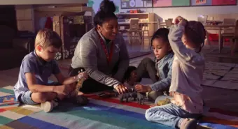 A child care educator sits on a carpet and plays with 3 children.