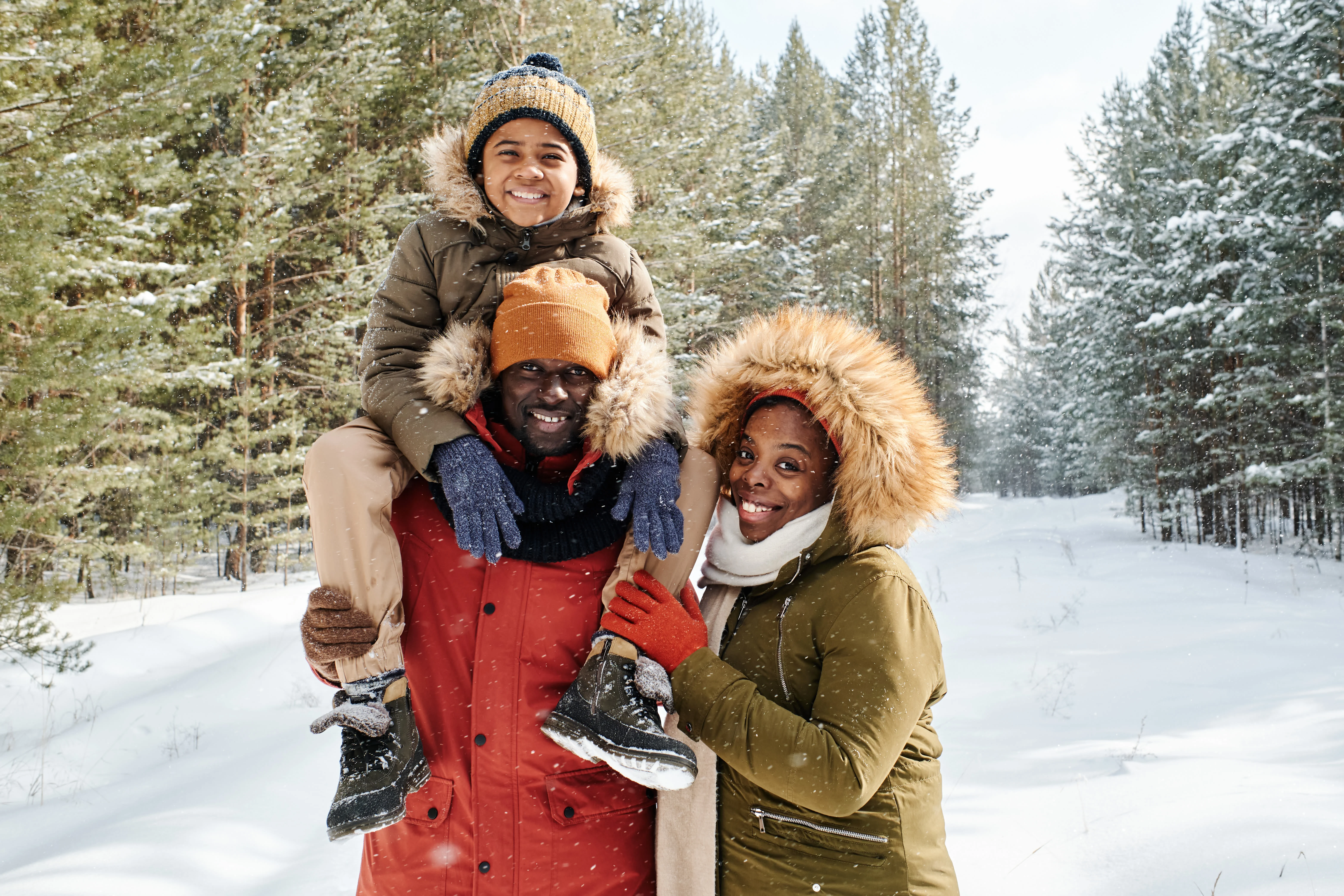 Family of three standing outside in snow wearing winter clothes. Child is sitting on one of the adults shoulders.