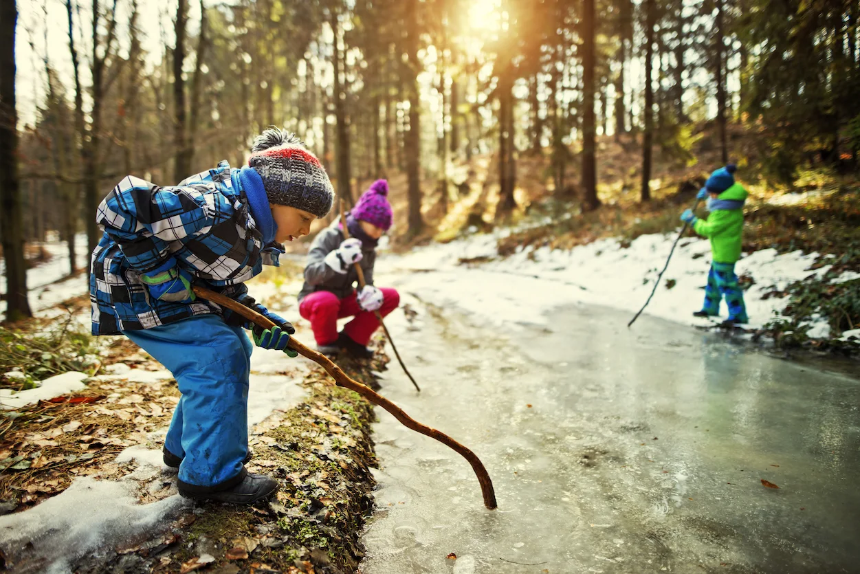 Three kids aged 10 and 6 are playing with a huge frozen puddle in winter forest. They are breaking ice on the puddle with sticks