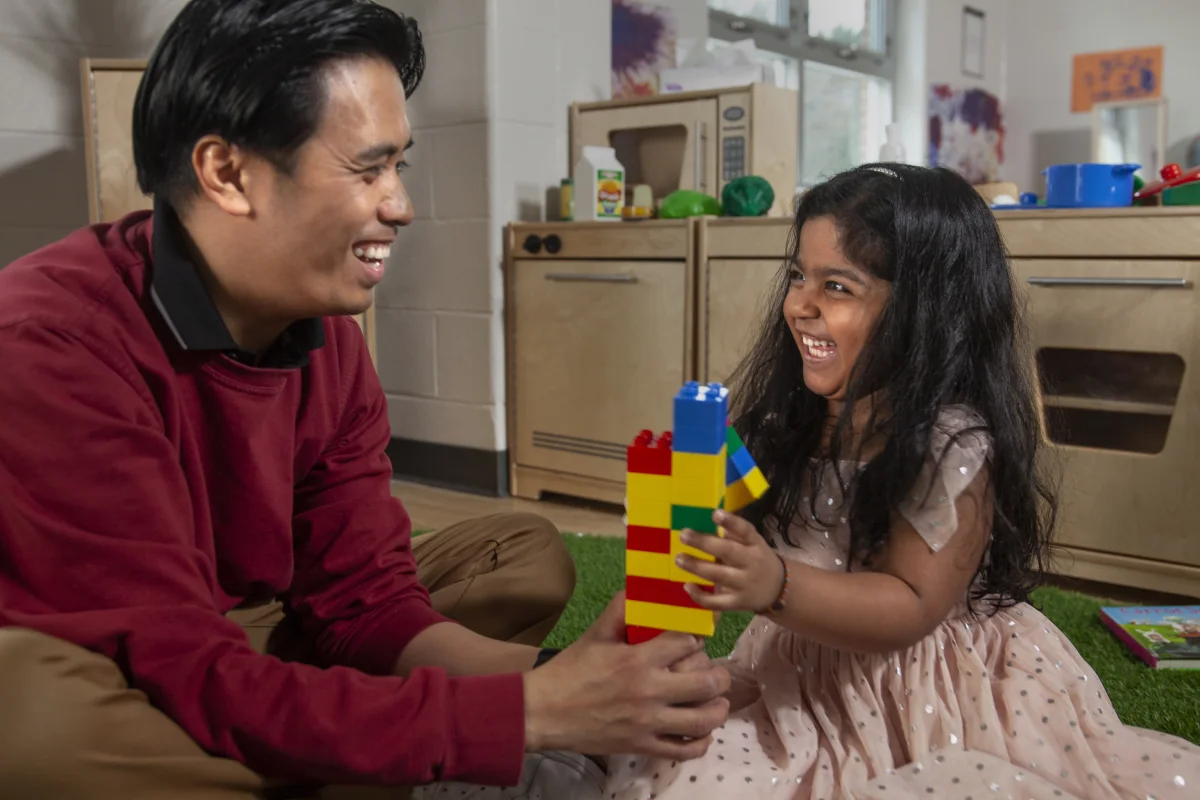Child care staff holding lego tower while preschool girl stacks on lego pieces